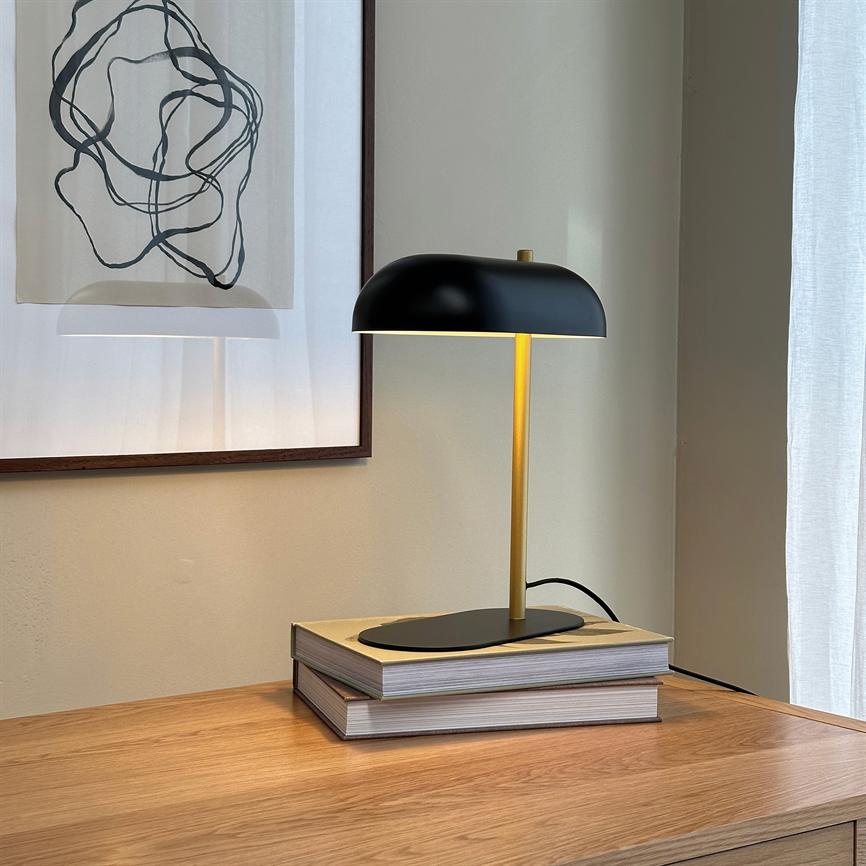 En sort og guldbelagt bordlampe står og kaster blødt lys over to stakkede bøger på et lyst træbord foran en indrammet abstrakt tegning og et hvidt gardin.