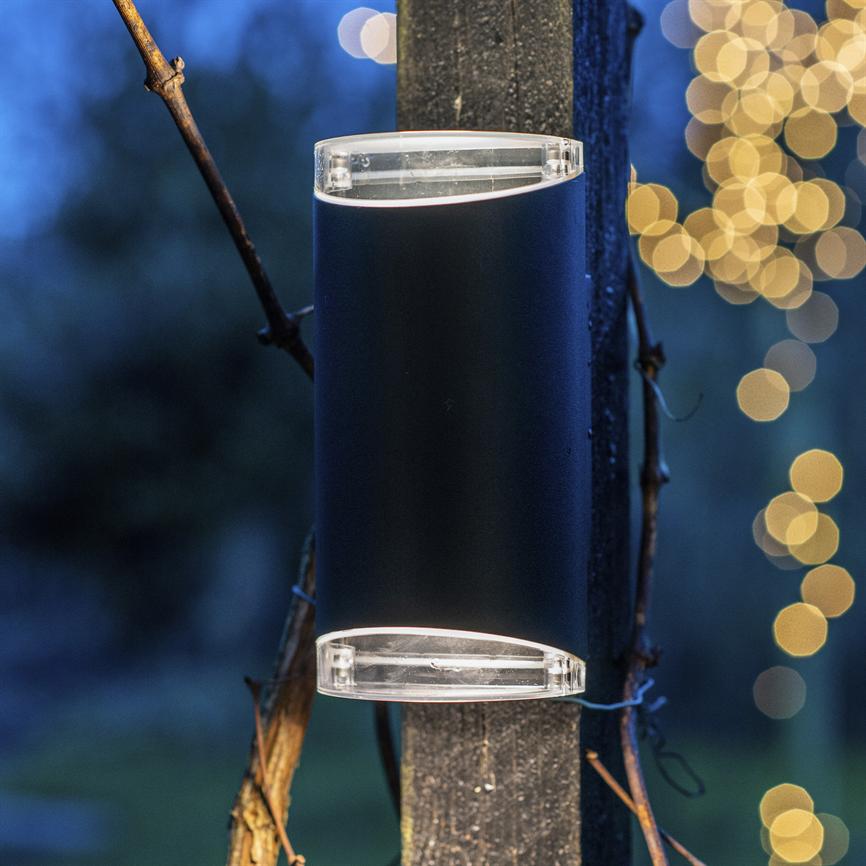 Outdoor lamp mounted on a wooden post, casts light upwards and downwards; the surroundings are evening blue with creepers around the post and warm, fuzzy yellow string lights in the background.