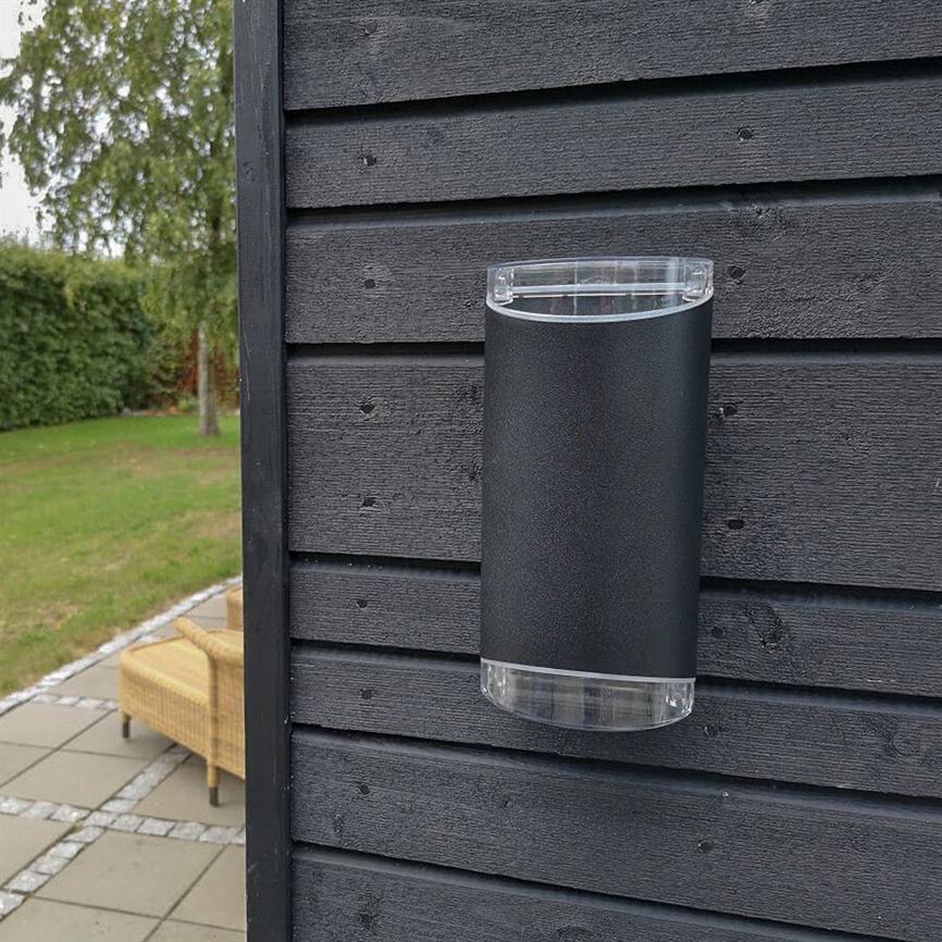 Wall lamp, cylindrical, mounted on black wooden cladding, does not light; clear plastic at the top and bottom. In the background lawn, hedge and wicker garden chair by the tiled terrace.