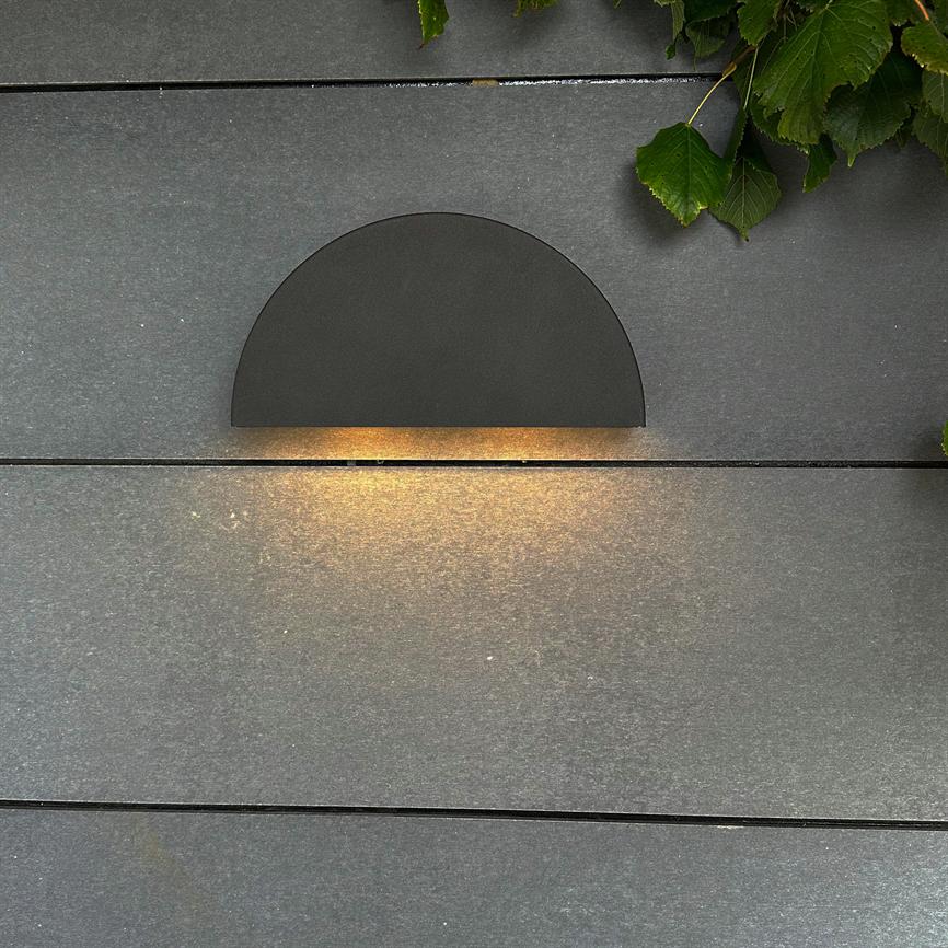 Semi-circular wall lamp emits a warm, downward glow on dark, horizontal wooden boards; green leaves poke in from the upper right corner suggesting an outdoor environment.