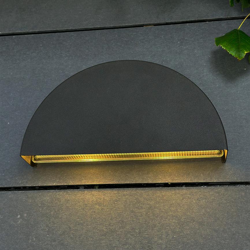 Semi-circular black wall lamp emits warm, golden light from a narrow opening, mounted on dark wooden planks with a green leaf in the upper right corner.
