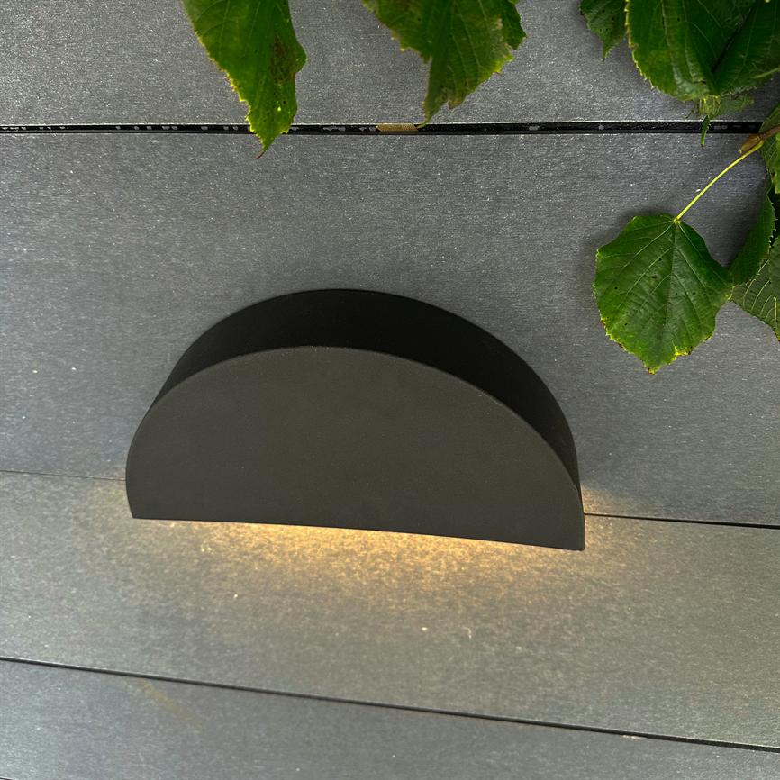 Black semi-circular wall lamp emits warm light downwards on dark wooden panels, with green foliage visible in the upper right corner.