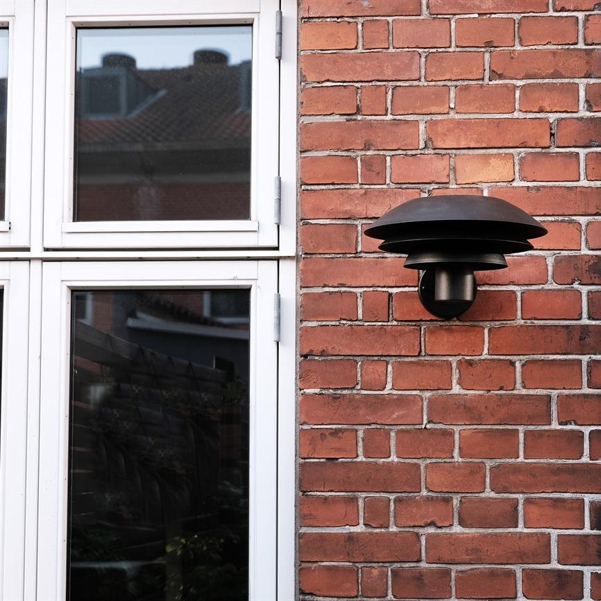 Black, bowl-shaped wall lamp mounted on red brick wall by a white-framed window; the window reflects roofs and surrounding buildings.