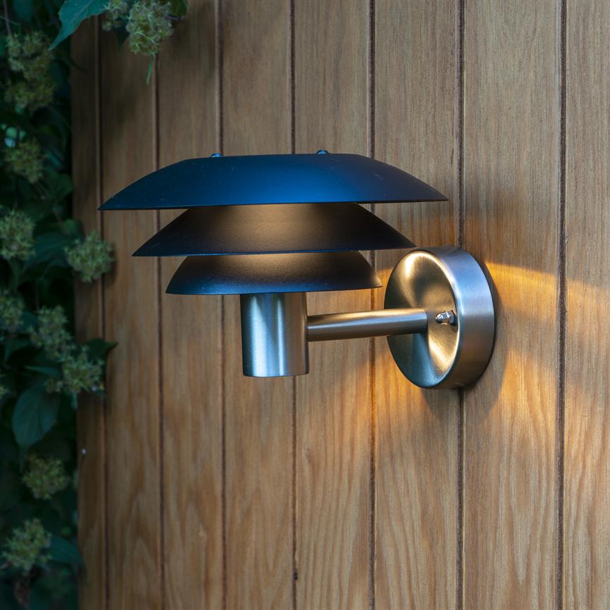 Brushed steel wall lamp with three shades shines softly downwards and spreads warm light on a vertical wood-paneled wall; mounting brackets and green foliage can be seen on the left.