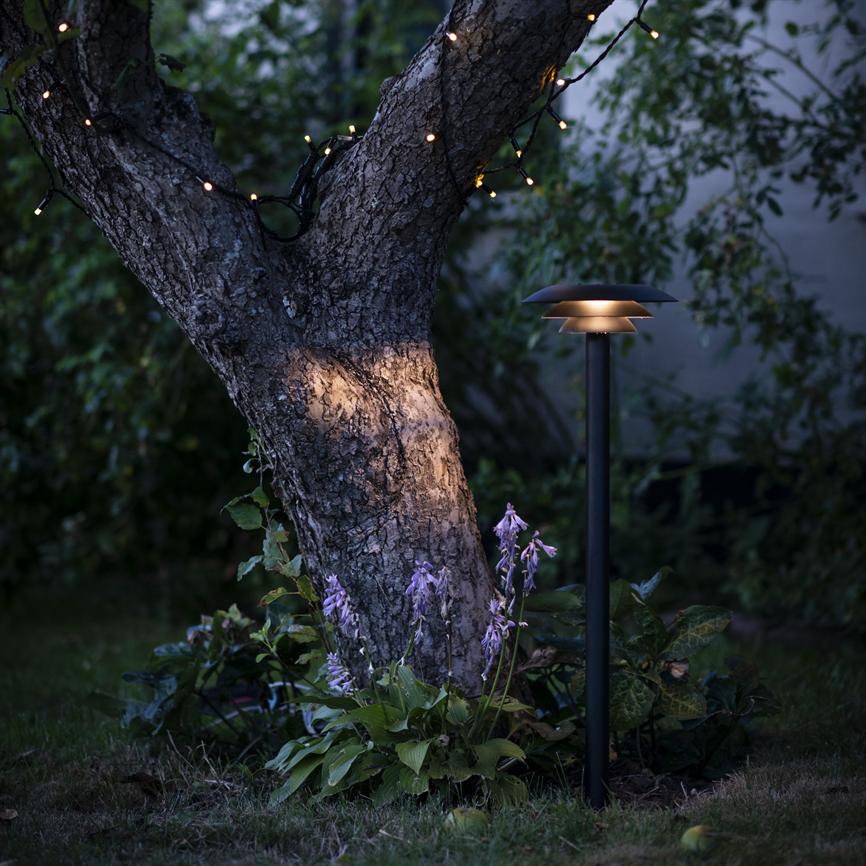 Tree trunk is illuminated by a low black garden lamp and small festive string lights, surrounded by purple hosta flowers and dense, dark garden foliage in the evening twilight.