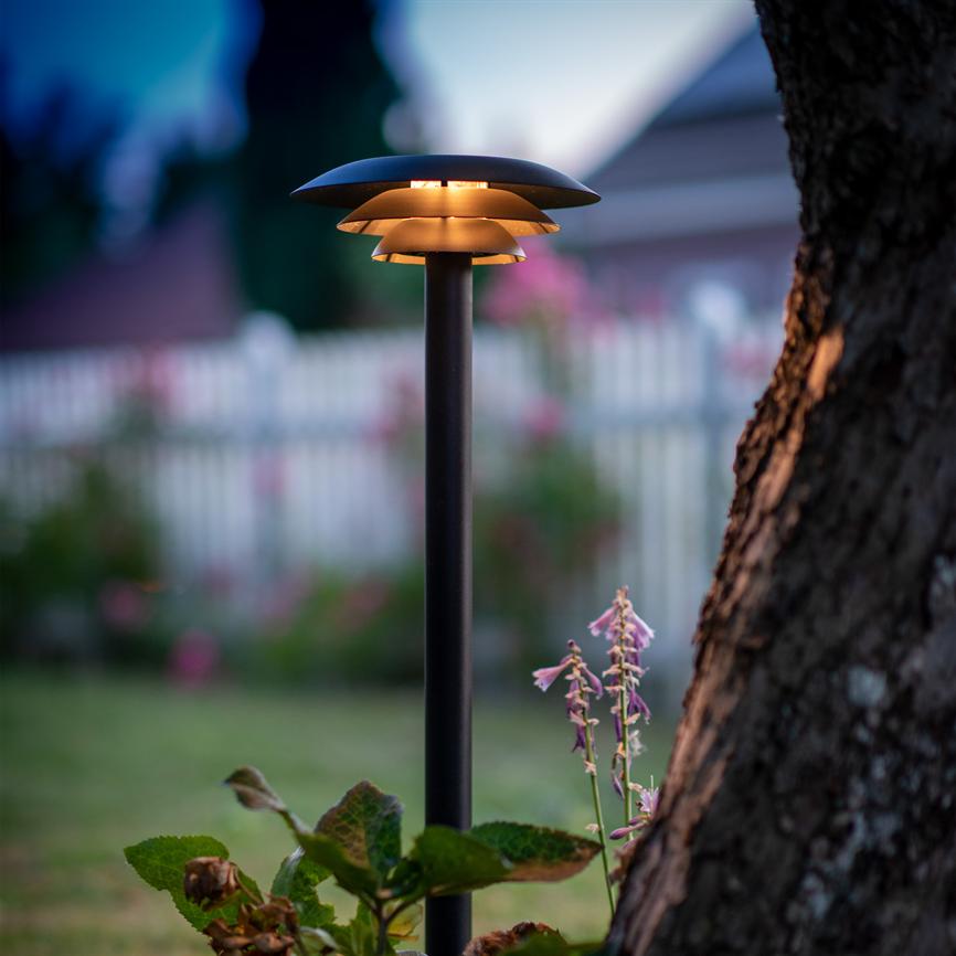 A black garden lighting lamp emits warm, downward light, standing by a tree trunk and flowers in the front garden. Background: blurred white picket fence and evening clouds at twilight.