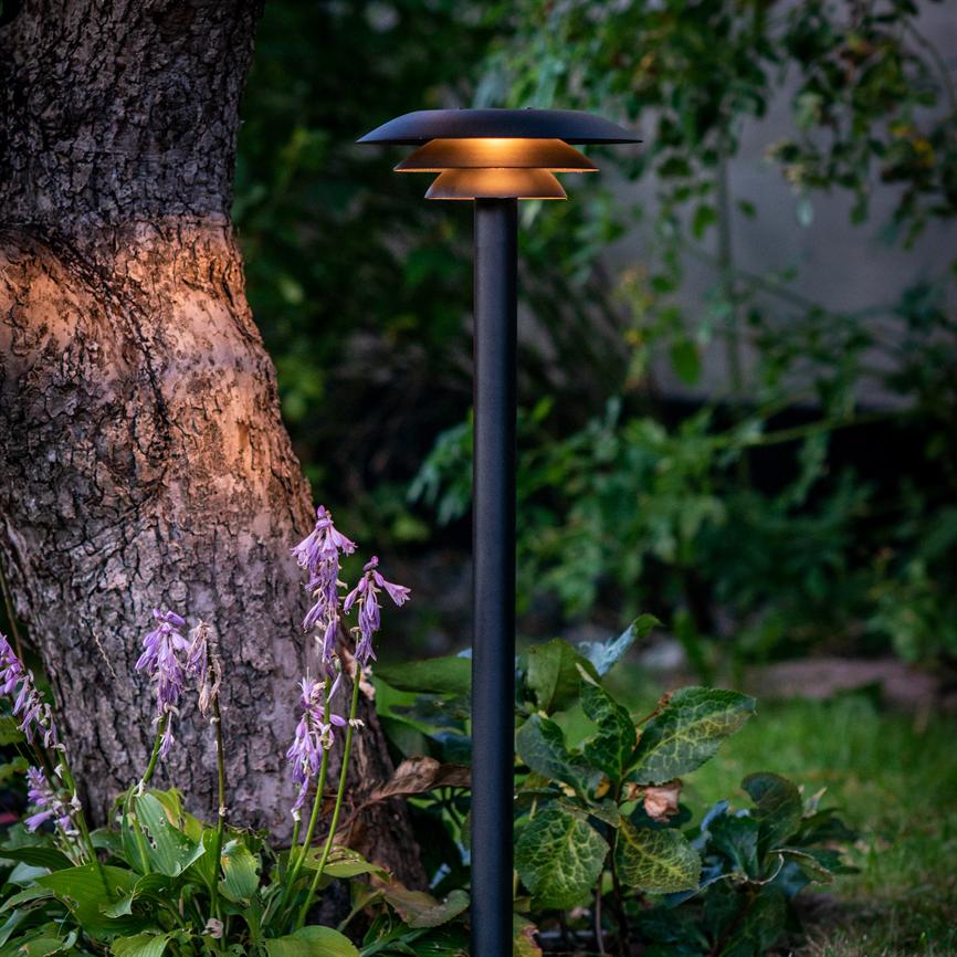 Garden lamp casts a warm, diffused glow downwards; it stands by a thick tree trunk among purple flowers and green leaves in a subdued, lush garden.