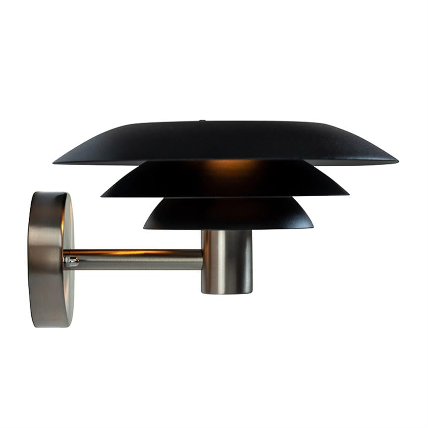Wall lamp with three layers of black shades mounted on a brushed metal arm, shines weakly downwards and casts a warm glow against a neutral, white background.