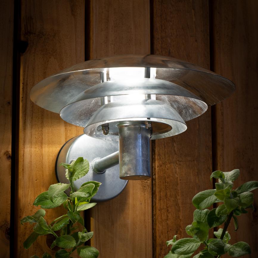 Silver metal wall lamp shines brightly downwards and outwards, mounted on vertical wooden panels; green plants under the lamp are caught by the light.