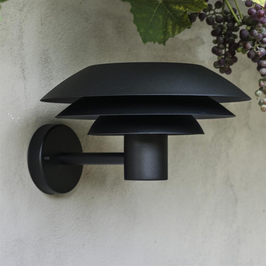 Black modern wall lamp with three layered shades, mounted on a light plastered wall; arm emanating from a round plate, with bunches of grapes and green leaves at upper right.