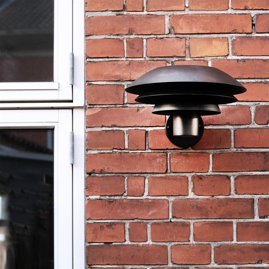 Wall lamp is mounted on a red brick facade; dark multi-layered round metal casing, placed next to a white window frame in an outdoor townhouse environment.