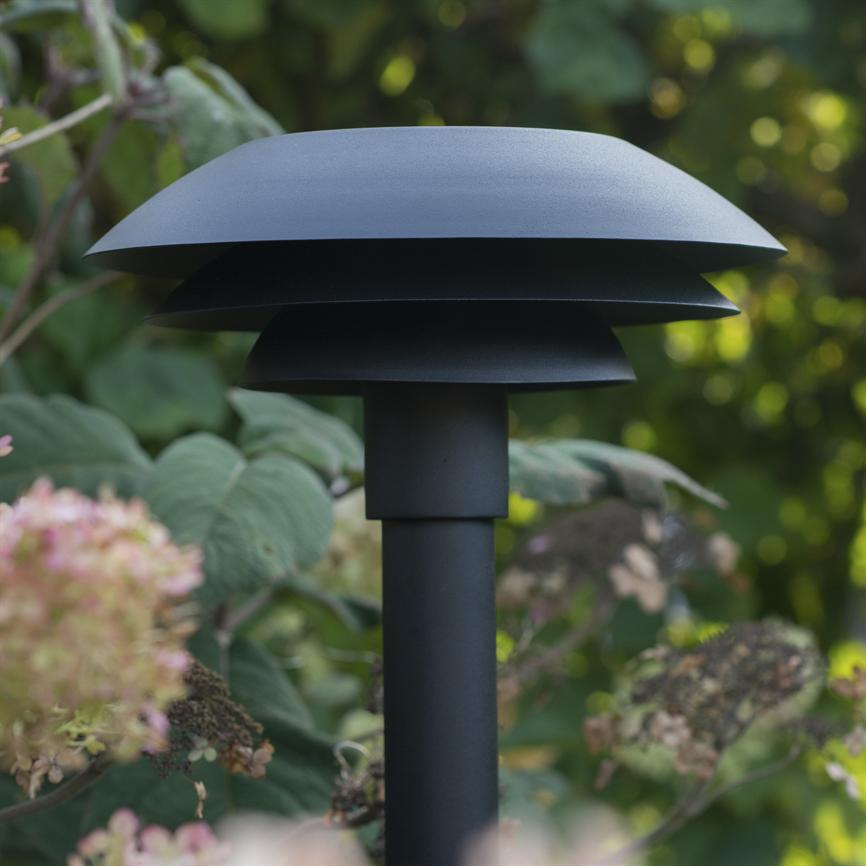 Black garden lighting lamp stands in a lush garden, with flat, tiered shades and a vertical post surrounded by green leaves and wilted hydrangea flowers.
