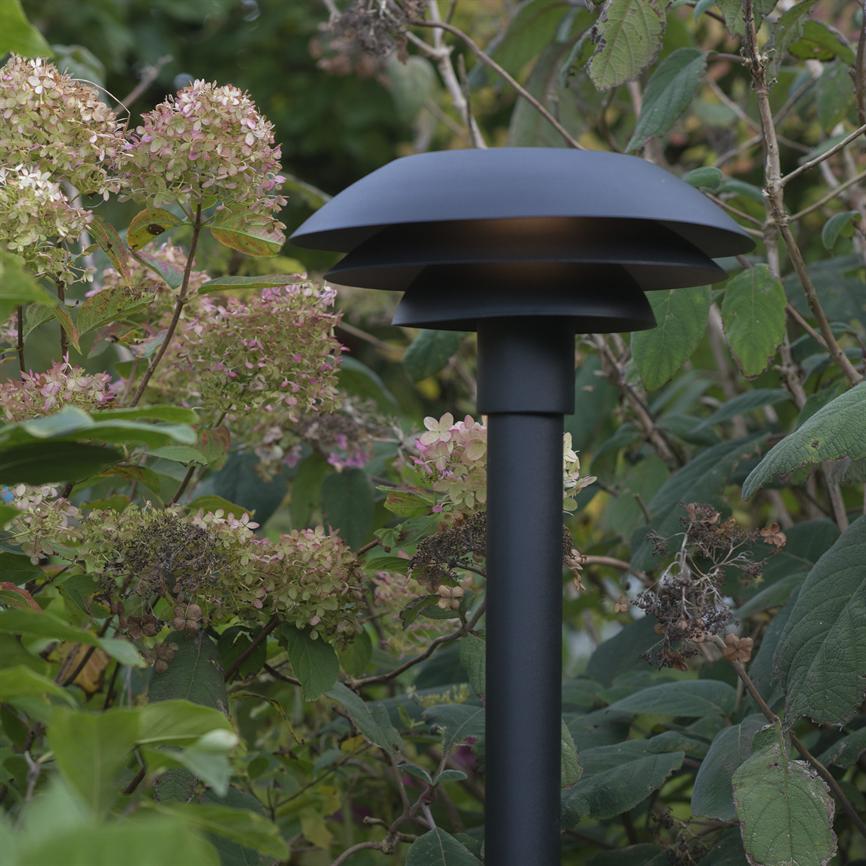 Mushroom-shaped black garden lamp stands among hydrangeas and shrubs, emitting weak downward light through layered shades in a lush, green garden.