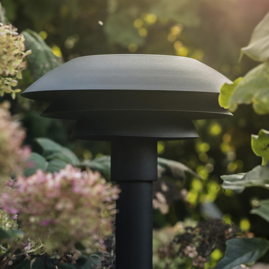 A black, mushroom-like garden lighting lamp stands on a thick column, placed among green leaves and pinkish-white flowers in a sunlit garden background.