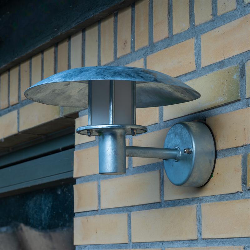 Wall lamp in galvanized metal mounted on a yellow brick facade; round shade pours over a cylindrical arm and lamp housing, placed close to a dark windowsill.