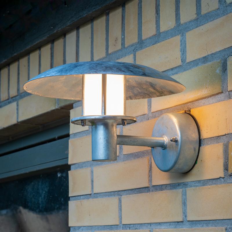 A metal wall lamp emits warm, soft light; it is mounted on a yellow brick wall with a round wall panel and disc-shaped screen, located by a door or window opening.