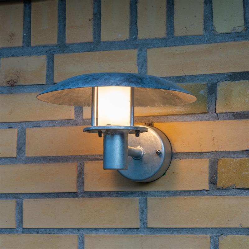 Outdoor wall lamp with round, screen-shaped shade glows softly; mounted on round metal plate attached to yellow brick wall with clear joints.