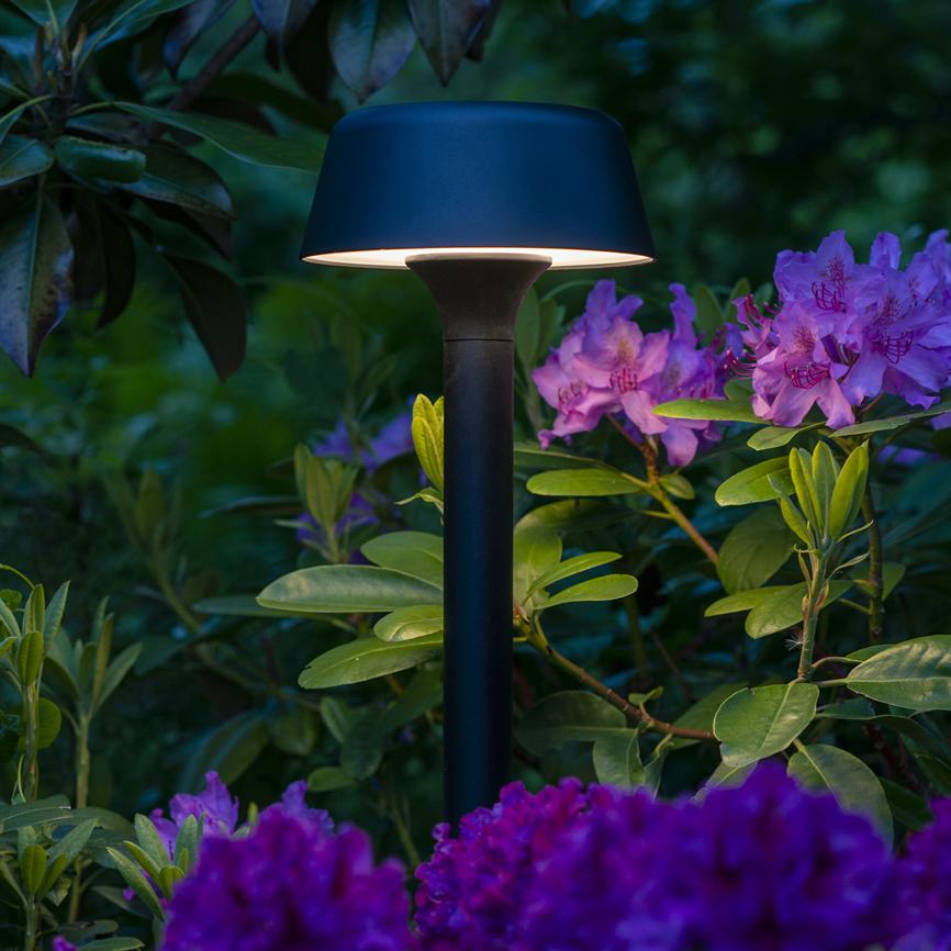 Black, mushroom-like garden fur lamp glows softly downwards; surrounded by green leaves and light-purple rhododendron flowers in a lush, subdued evening garden.