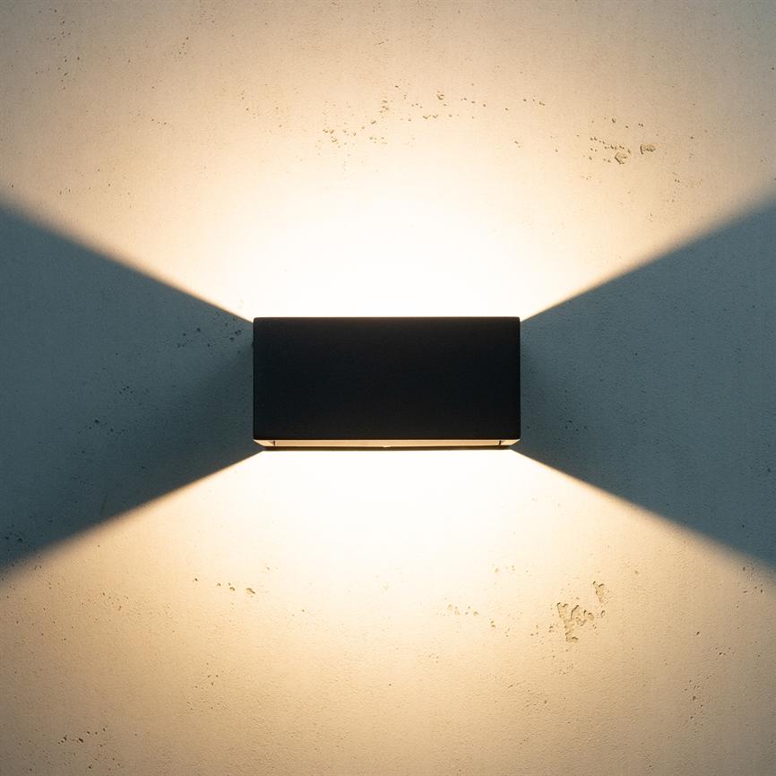 A black rectangular wall lamp emits warm cone-shaped beams of light upwards and downwards, projecting symmetrical triangular shadows on a light, lightly textured wall.