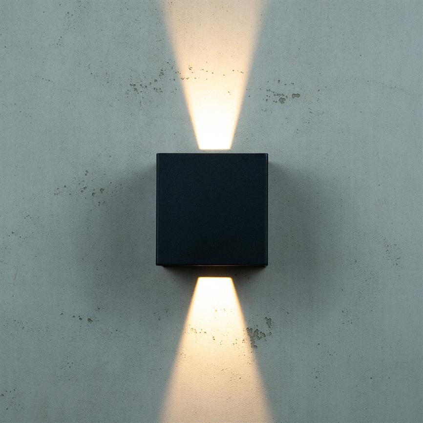 Black square wall lamp emits two cone-shaped beams of light upwards and downwards towards a grey, slightly patinated concrete wall.