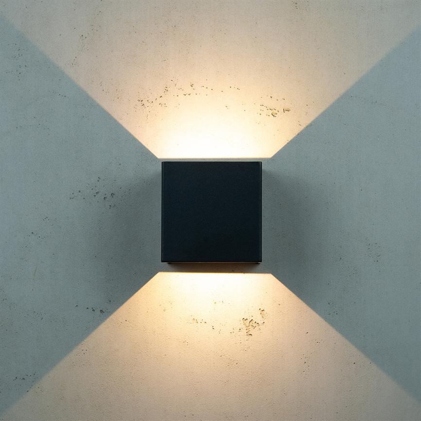 Black, square wall lamp mounted centrally emits warm light directed upwards and downwards, forming cross-shaped beams of light on a light grey, slightly rough concrete wall.