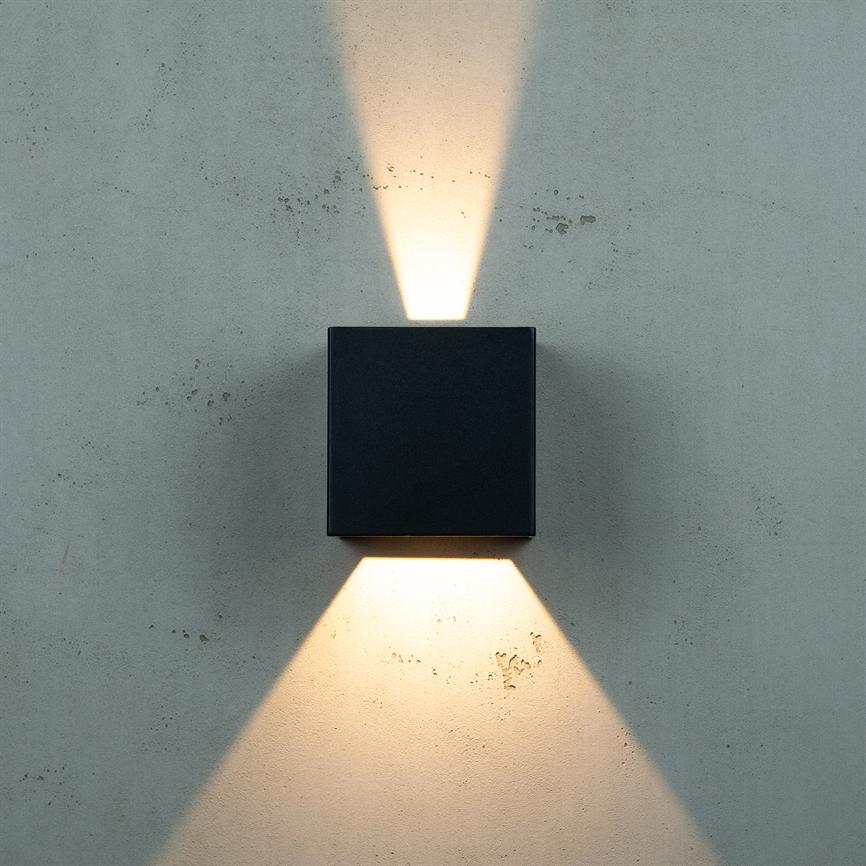 A black square wall lamp emits warm light rays upwards and downwards, forming triangular patterns on a raw, gray concrete wall.