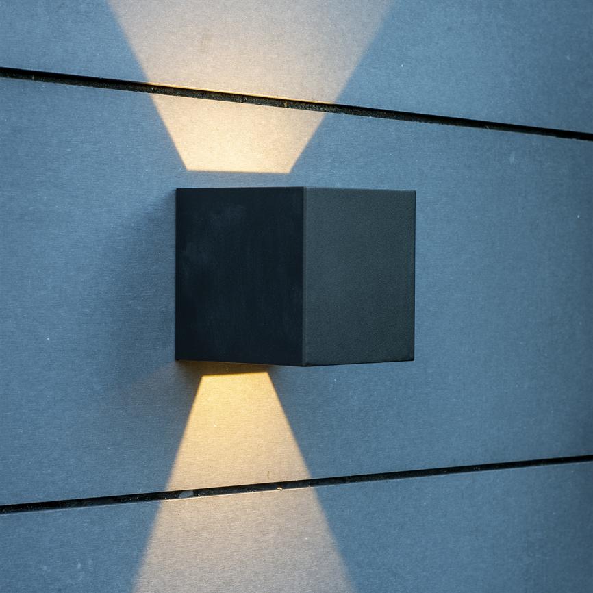 A black, cube-shaped wall lamp sends narrow, warm cones of light up and down against a blue-grey paneled wall with clear horizontal joints.