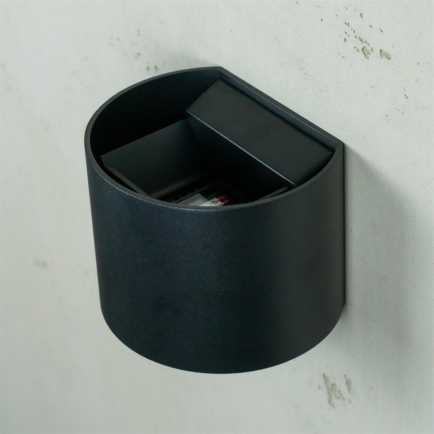 Black cylindrical wall mounted container with open top; interior compartment and small partition, holds a small object, mounted on a light, slightly dirty wall.