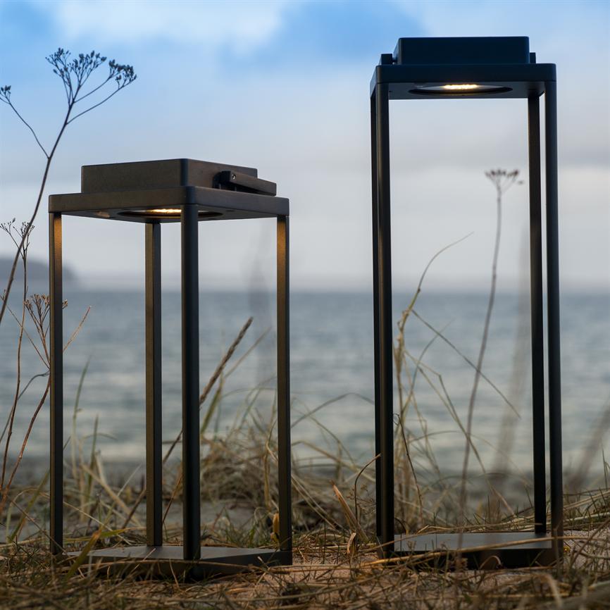 Two black, rectangular metal lanterns stand dimly lit on a sand dune with dry straw and wild flowers, in front of a calm sea under a bright, cloudy sky.