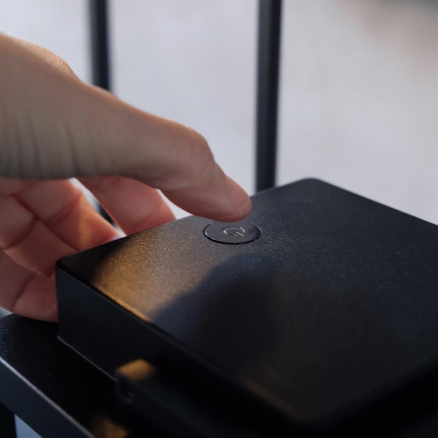 Hand presses the on/off button of the box with a fingertip; the box is black and rests on a piece of furniture with dark metal bars in a blurred background.