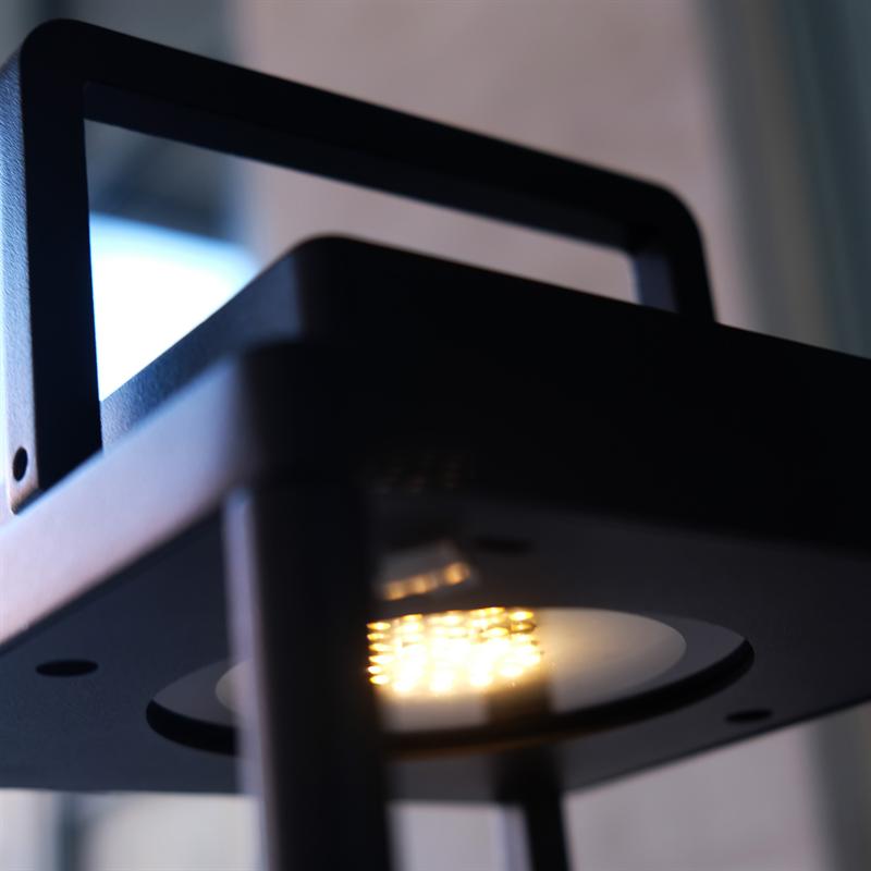 Black lamp body with visible LEDs emits warm yellow light, mounted in a metal frame in front of an obscured window.