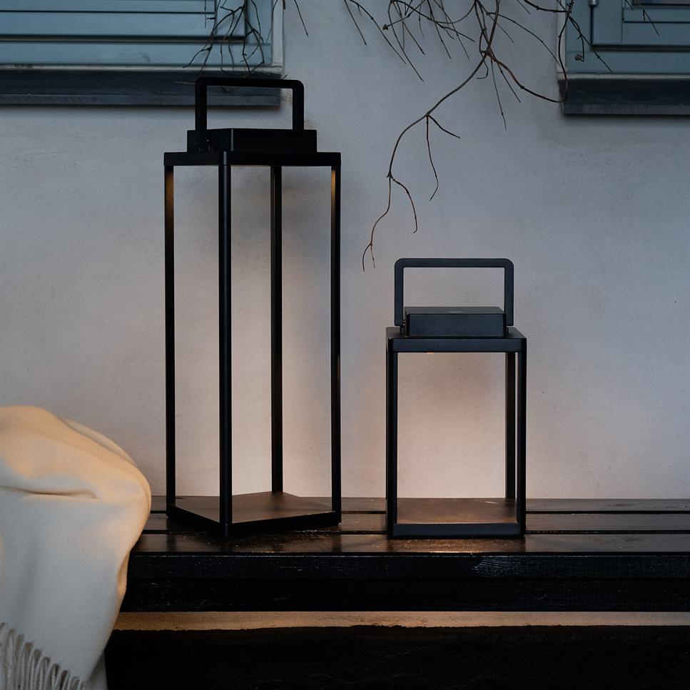 Two black rectangular lanterns stand on a dark bench, both emitting warm light; light gray wall, window sill and bare branches behind them, with light carpet hanging to left.