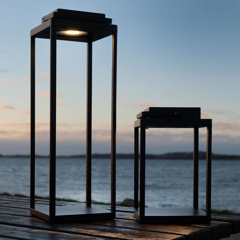 Two black, rectangular lanterns stand on a wind-worn wooden deck; one is on and casts a dim light, the other is off, in front of calm sea and pastel twilight sky.
