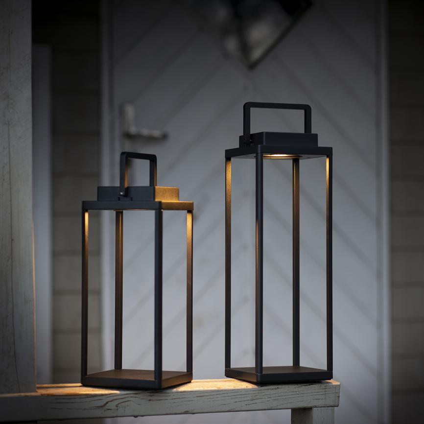 Two black metal lanterns at different heights radiate warm, subdued light; they stand on a worn white railing in front of a door with slanted panels in a twilight mood.