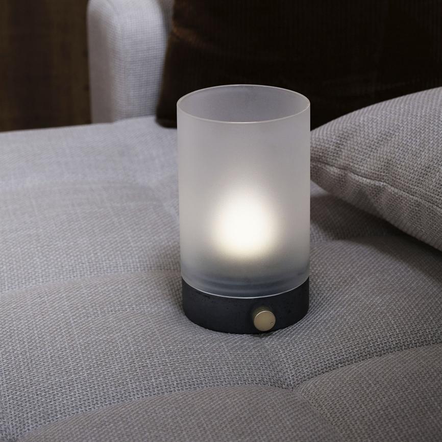 A small cylindrical lamp glows dimly from within; it stands on a light gray fabric sofa next to a light gray pillow in a subdued, cozy living room.