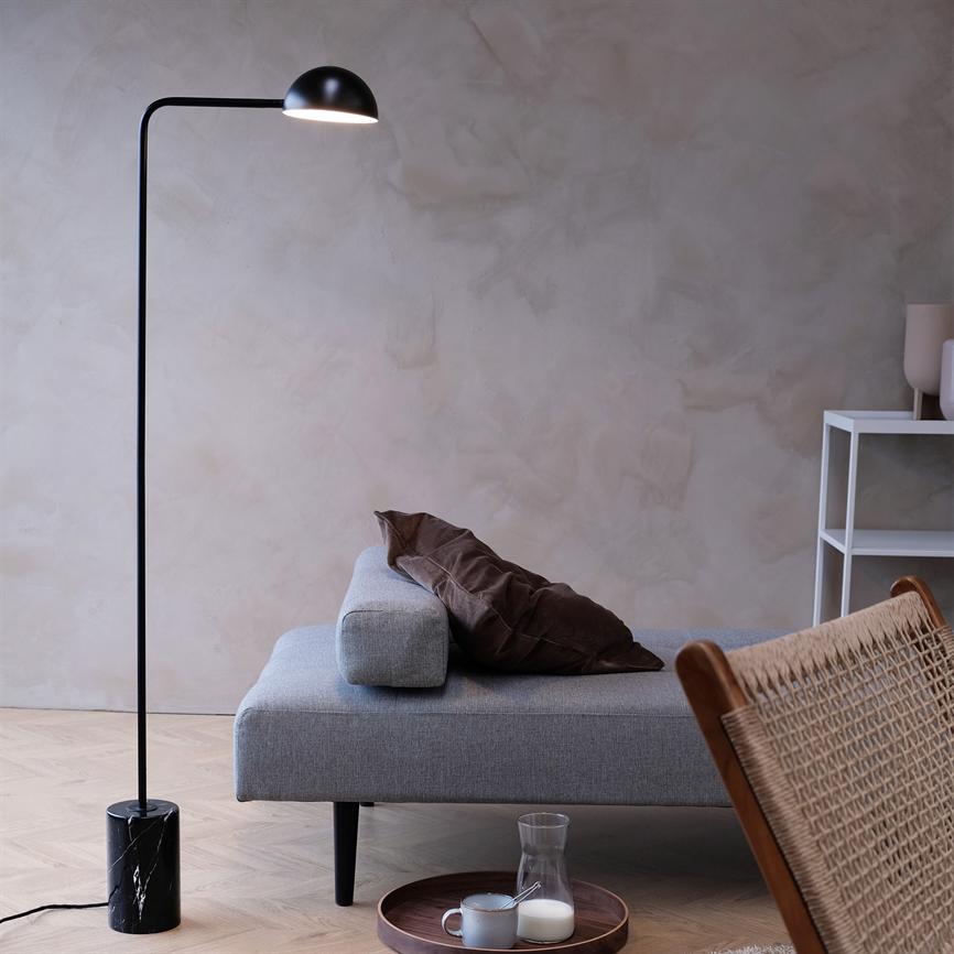 Black floor lamp shines down on a low gray sofa; a brown cushion cover lies on the sofa, and a round wooden dish with a cup and milk jug stands on a light wooden floor in a minimalist living room.