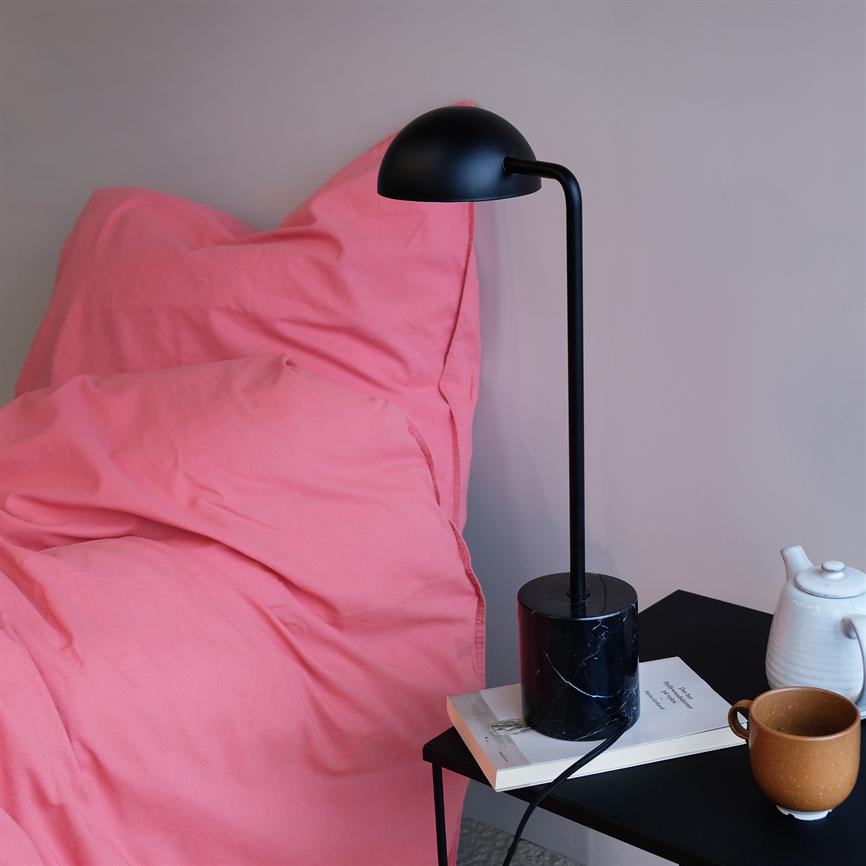 A black table lamp stands on a black bedside table next to a pink duvet; a cup, a teapot and a book lie next to it in a quiet bedroom.