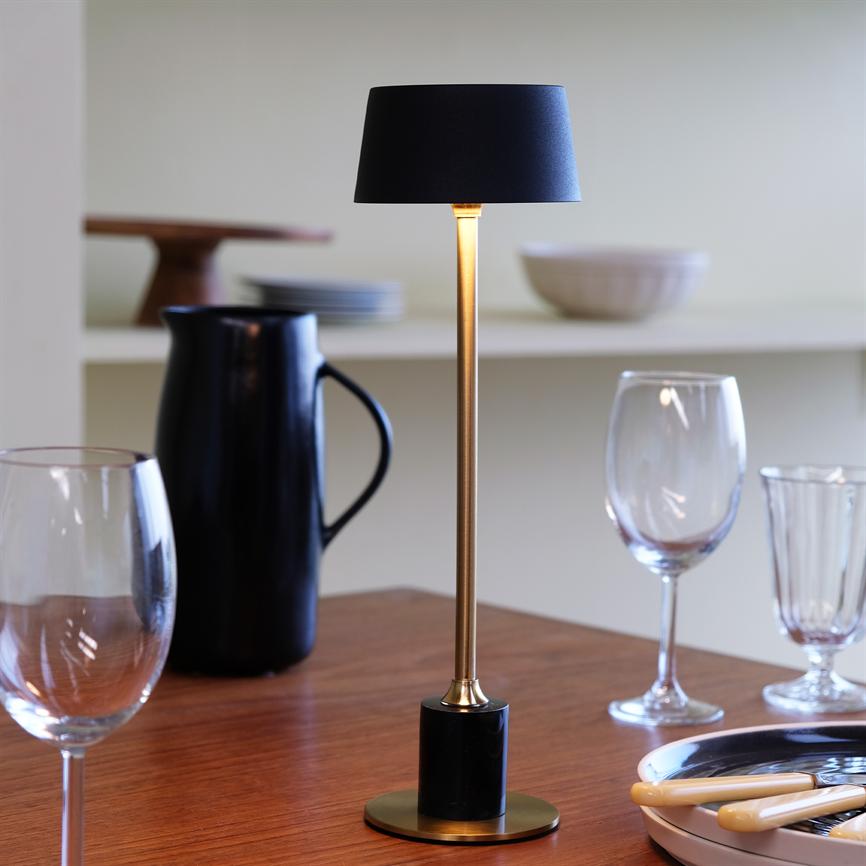 A slim table lamp is lit, casting a soft, warm light; it stands in the middle of a wooden table surrounded by wine glasses, cutlery, a black jug and dishes in a subdued dining room.