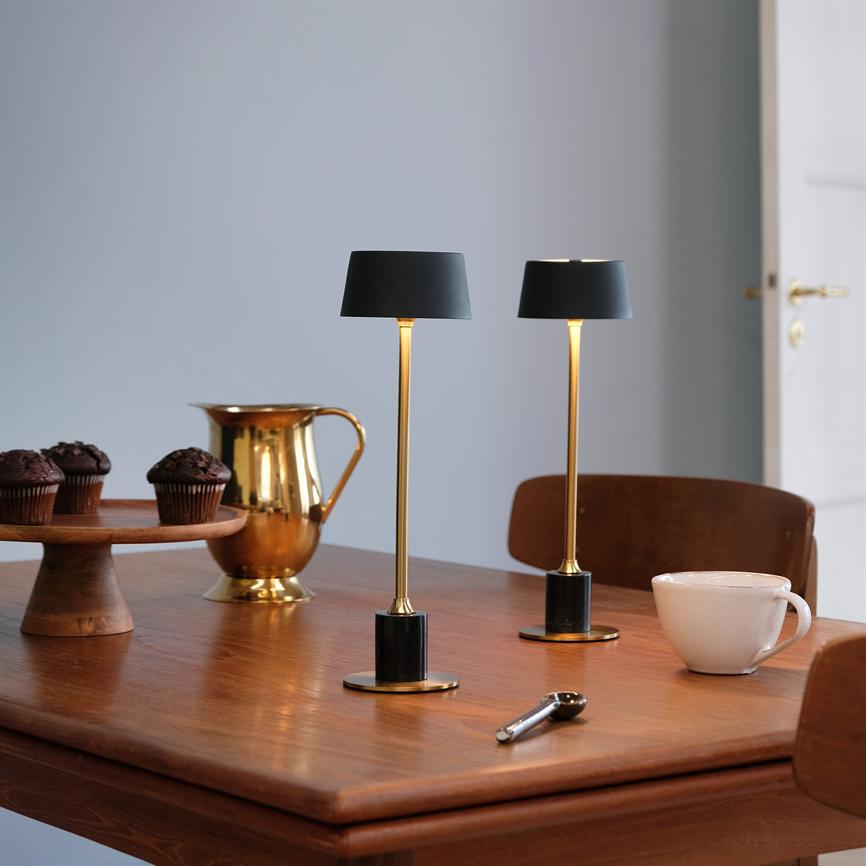 Two slim table lamps with black shades shine softly above a wooden table, surrounded by muffins on a cake plate, a golden jug, a white cup and a chair in a quiet room.