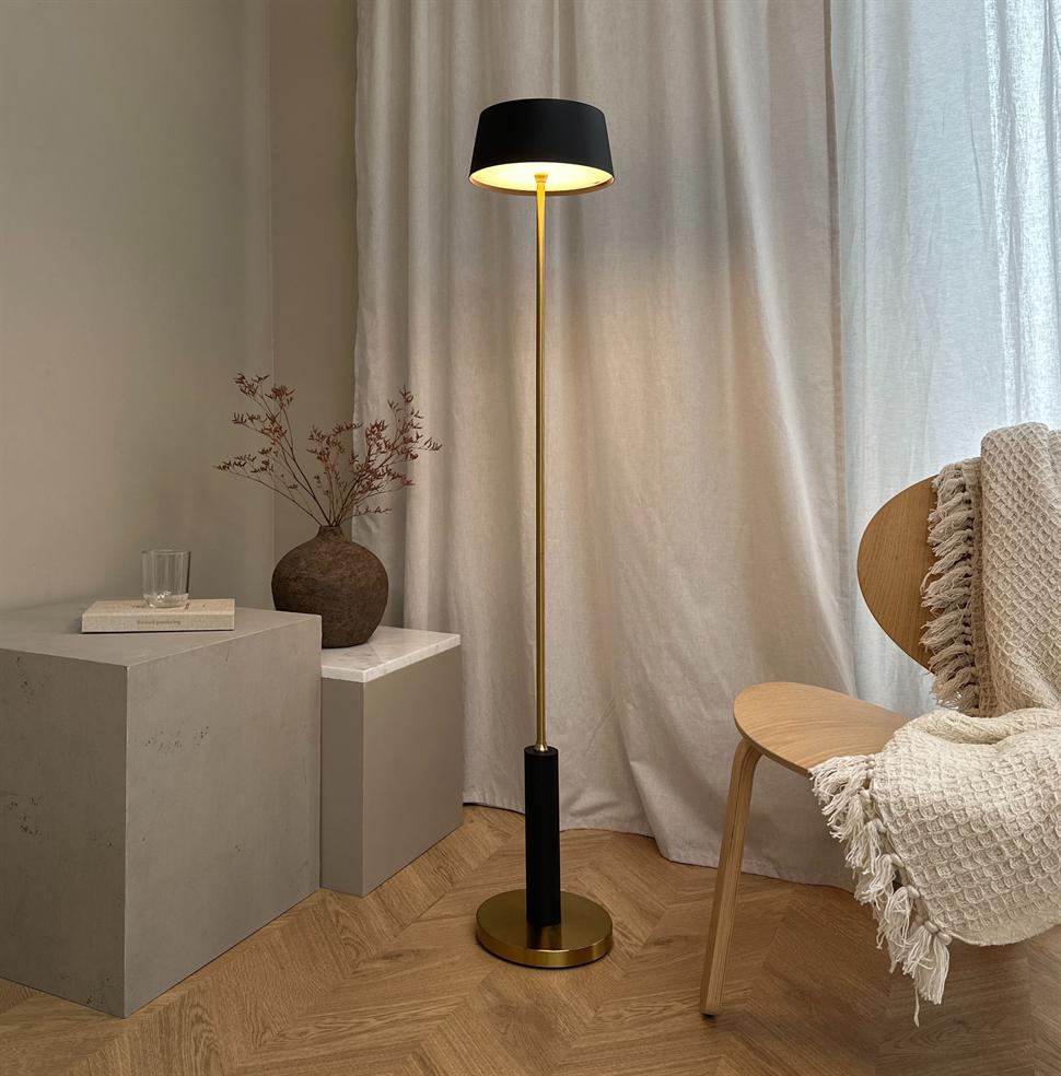 A slim floor lamp in black and brass is lit and casts a warm light; it stands by a wooden chair with a knitted plaid, in front of light curtains and a ceramic vase on concrete plinths.