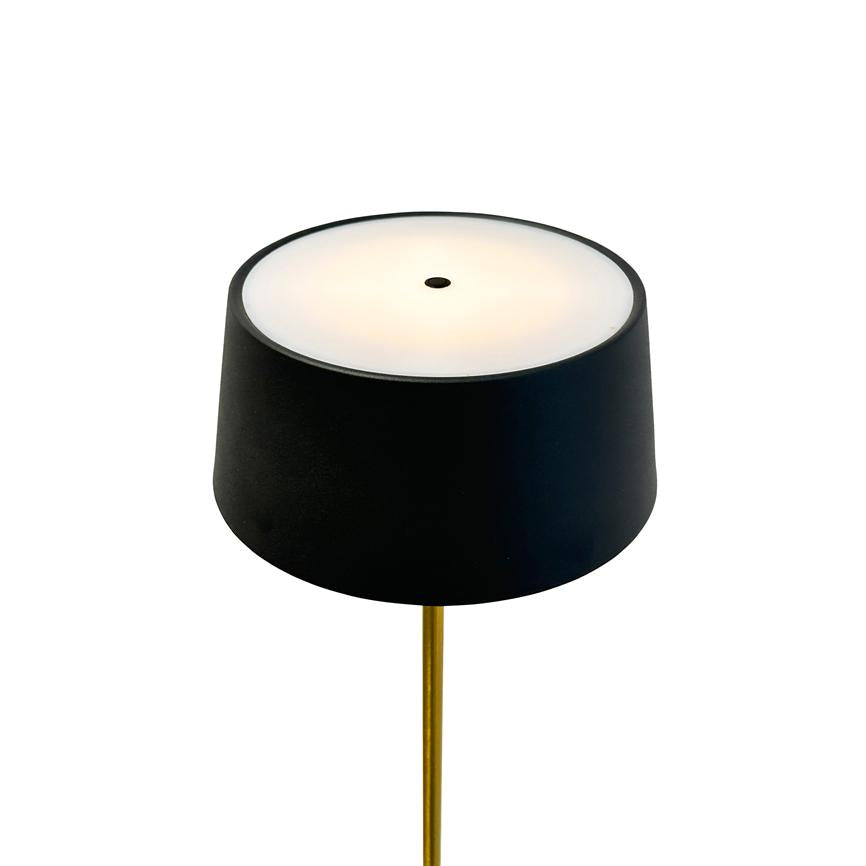 Black lampshade emits warm, diffused light through a white top with small central hole; mounted on a slender gold-plated rod against a white background.