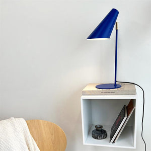 Blue lamp shines down on a book, placed on a white wall-hung box shelf; the shelf holds a small ceramic bowl and books, next to it a wooden chair with a white carpet.
 Text on the book: "new scandinavian design"