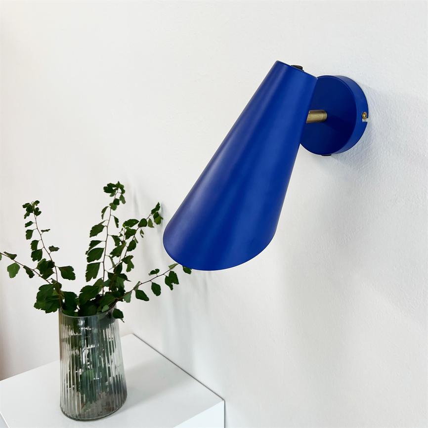 Blue conical wall lamp points down from a white wall above a white cabinet; a ribbed glass vase with green leaves stands on the cabinet.