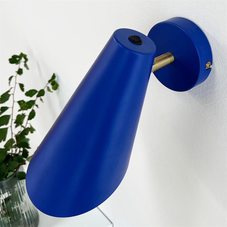 A blue cone-shaped wall lamp facing downwards, mounted on a round bracket with brass detail; white wall and a green plant in a glass vase can be seen in the background.