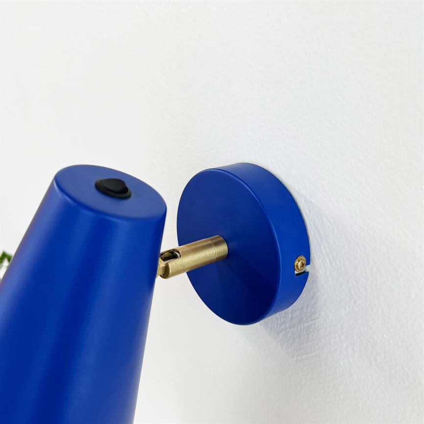 Blue wall lamp moves on a brass joint, with black switch on the lampshade; mounted on round blue wall plate, on a white wall.