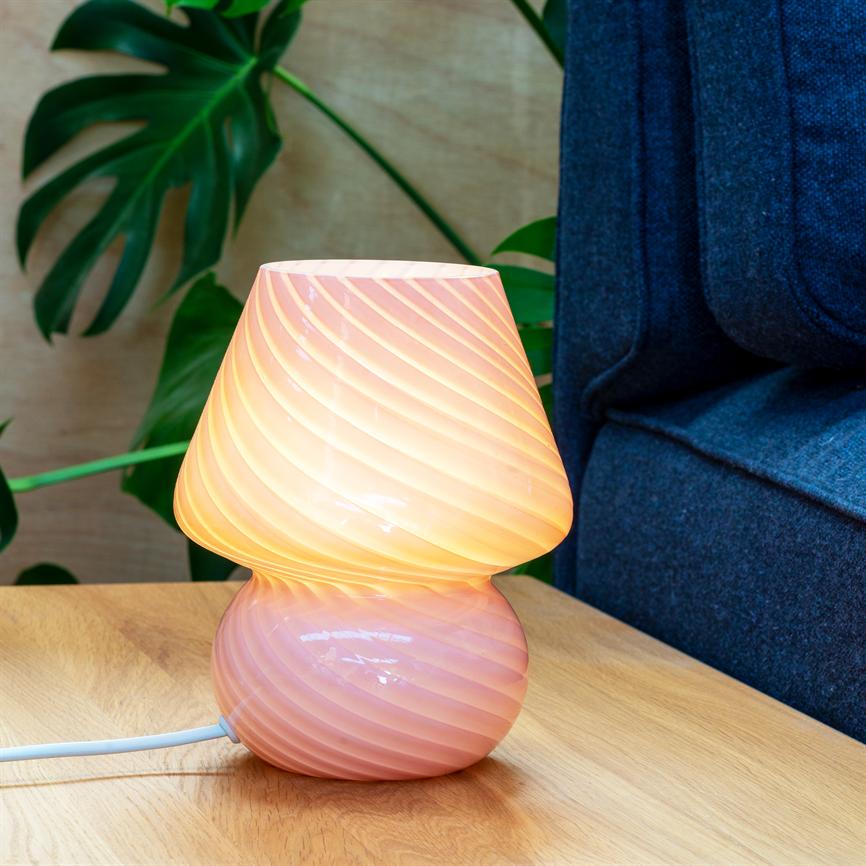 Lamp in pink glass glows with a warm, spiral-shaped glow; it stands on a light wooden table next to a dark blue sofa, with large green Monstera leaves in the background.
