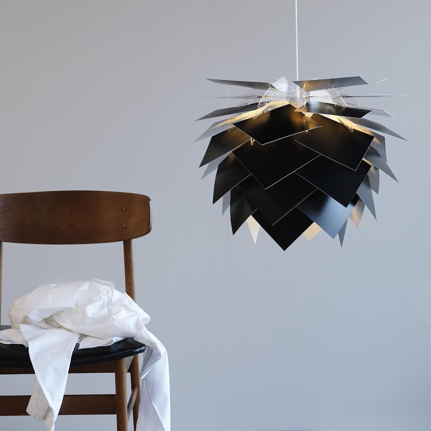 A tiered black pendant light hovers lit in front of a gray wall; to the left is a wooden chair with a white shirt draped over the seat.