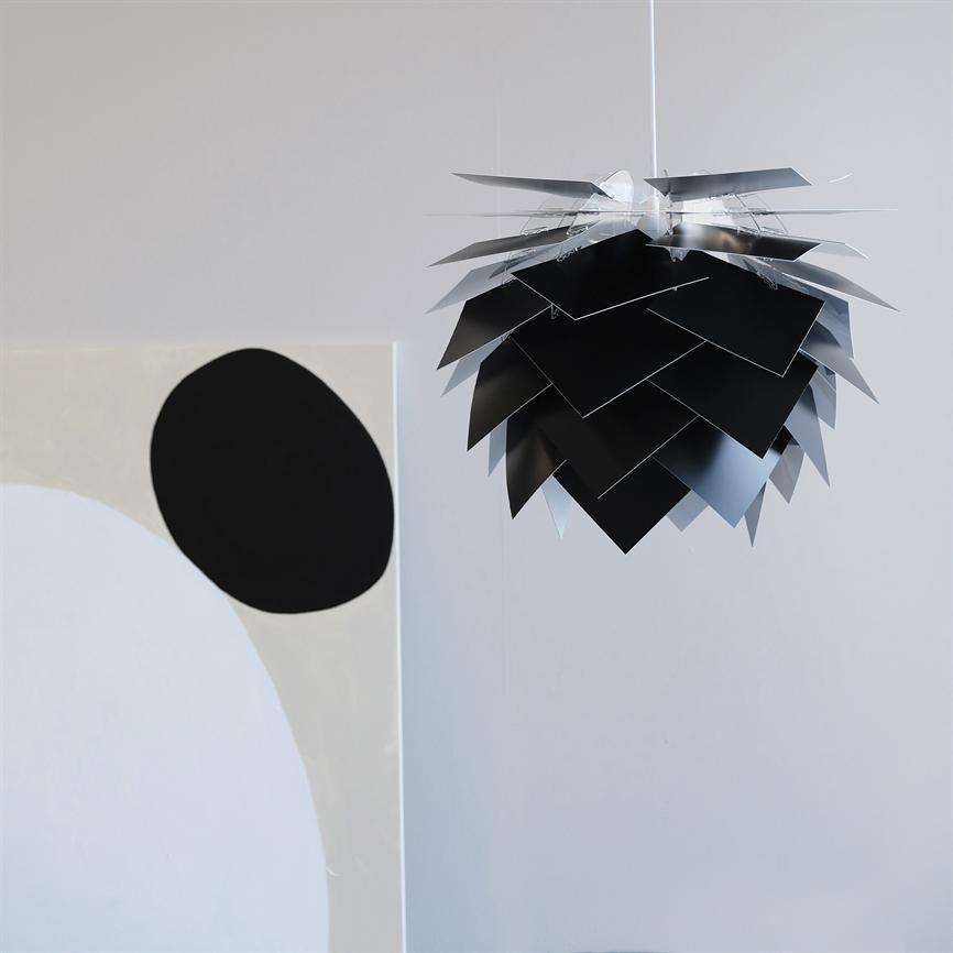 A black-and-silver tiered pendant light hangs from the ceiling; its overlapping metal plates form a cone-like shape. In a bright, minimalist room next to an abstract painting with a large black circle.