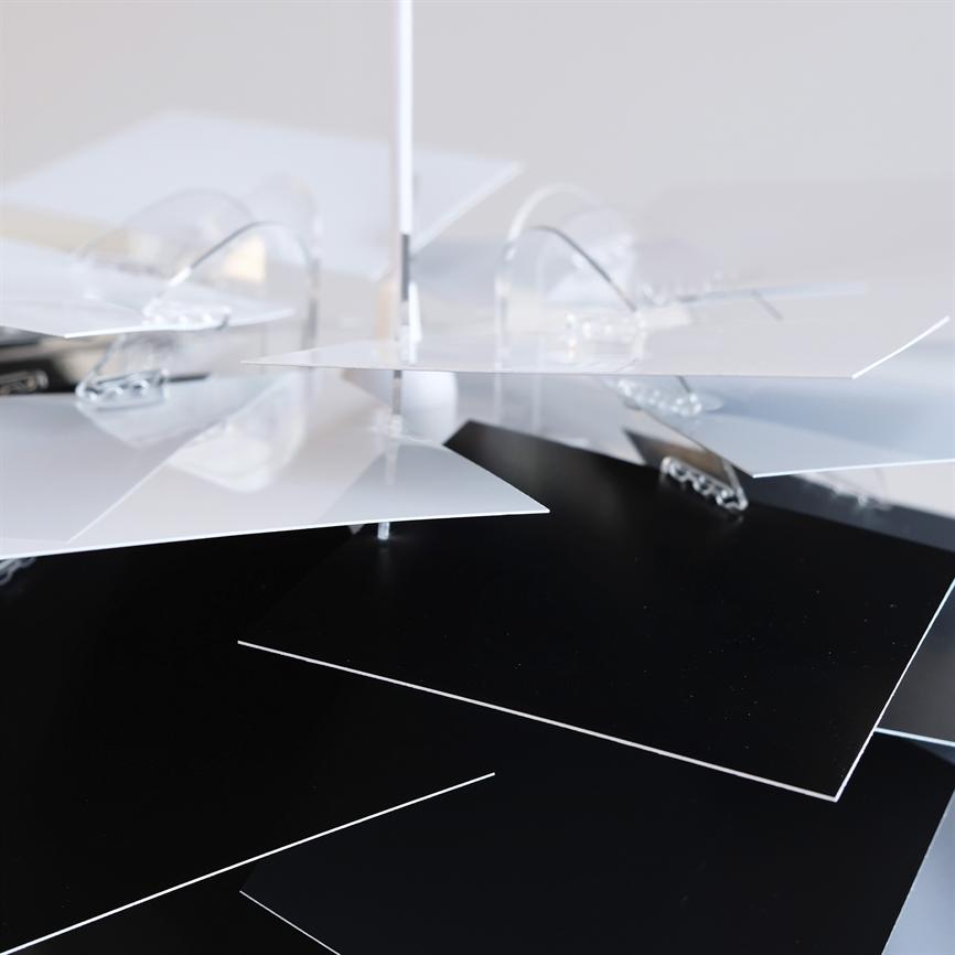 Black and white rectangular plates hang in overlapping layers from a vertical central bar; transparent round brackets hold them together. Scene in a bright, neutral environment.