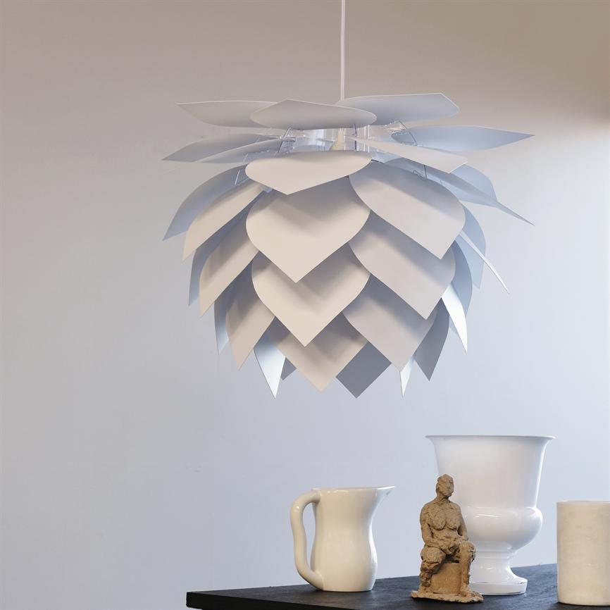 Hanging lampshade looks like layer-on-layer leaves; it hovers over a table. On the table is a white jug, a white vase and a small clay figure against a light, neutral wall.